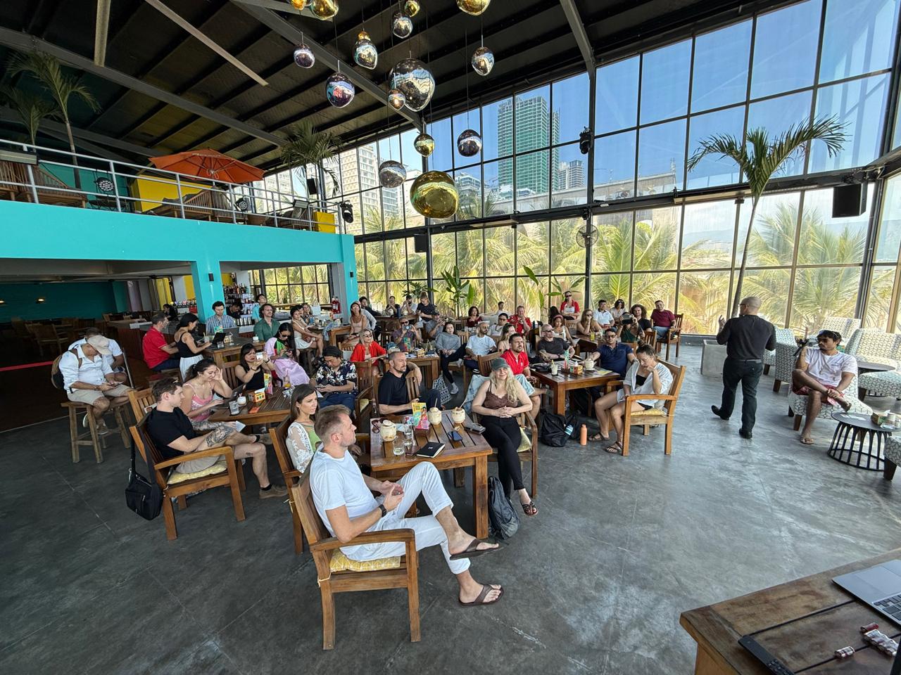 Full crowd at wooden tables, disco-ball chandeliers, panorama windows, presenter speaking.
